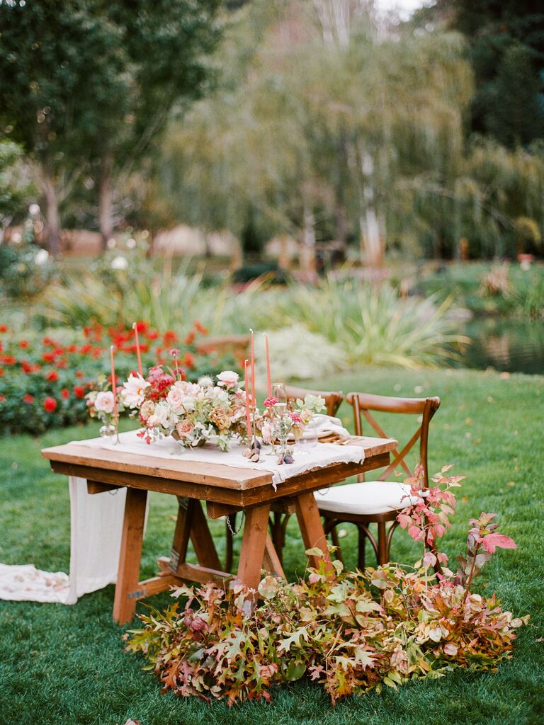 outdoor wedding sweetheart table decor with white gauze table runner, orange taper candles and fall foliage arrangements on the ground