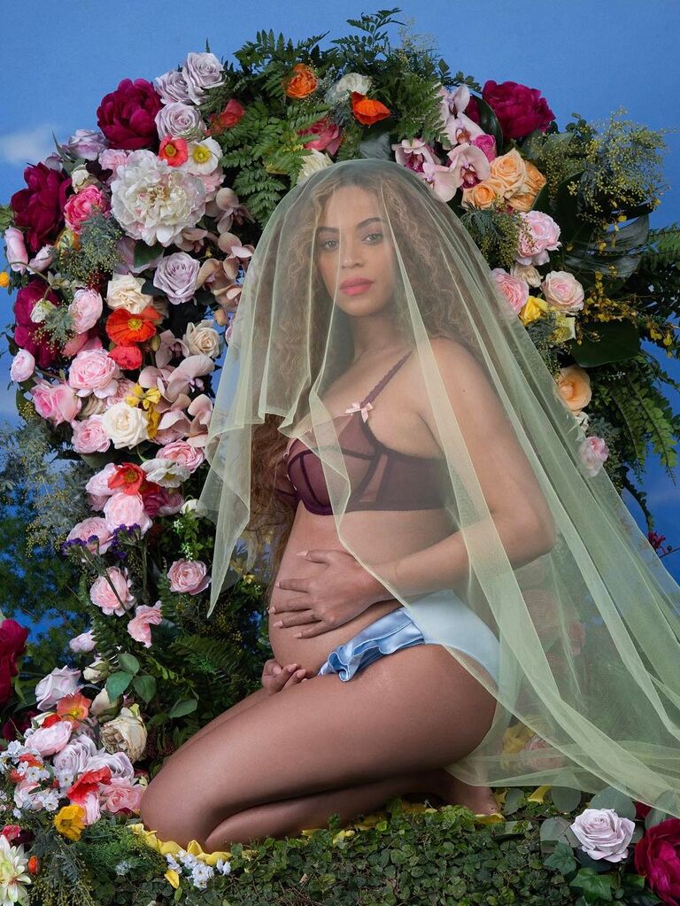 Beyonce poses for a maternity photo shoot with a lush floral background and a veil over her head. 