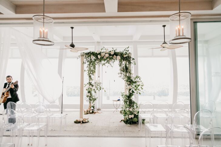 Minimal white chuppah with asymmetrical floral decorations surrounded by ghost chairs.