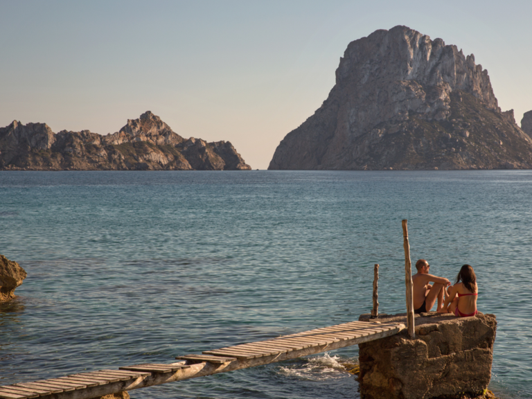 Tourist couple sitting on pier looking over to Es Vedra, Ibiza, Spain