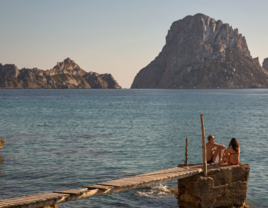 Tourist couple sitting on pier looking over to Es Vedra, Ibiza, Spain