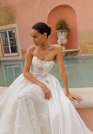 A-line silhouette dress made of Chantilly lace with delicate applications on the corset. The dress includes a detachable train, which is securely fastened with hidden buttons at the waist. The train is crafted from luxurious Mikado fabric and elegantly adorned with lace on top. The detachable train allows you to transform your look effortlessly, offering two styles in one dress. The gown features multiple layers of soft tulle lining. Perfect for brides seeking versatility and timeless elegance.