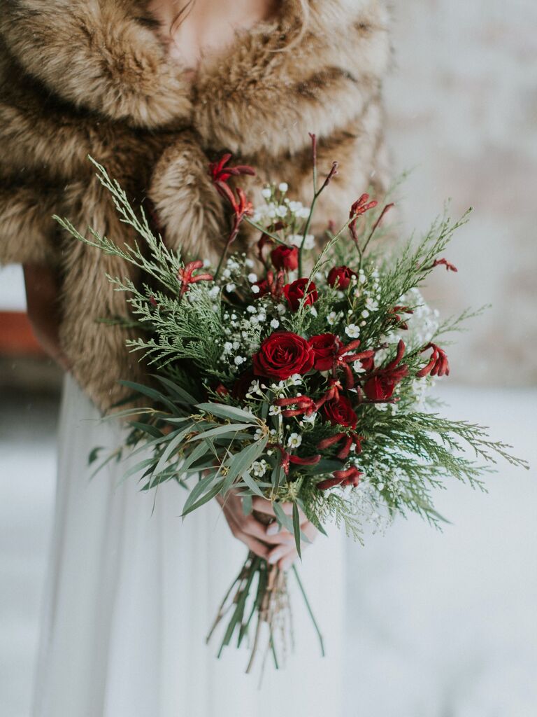 bride wearing brown fur wrap holds winter wedding bouquet with red spray roses and evergreen branches