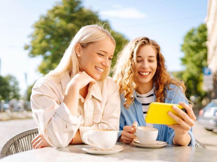 Best friends having coffee outside while smiling at a phone