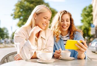 Best friends having coffee outside while smiling at a phone