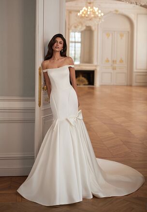The Moonlight Collection J7006 wedding dress is a sleek satin mermaid gown featuring an off-the-shoulder neckline, sculpting princess seams, and a soft basque waist for a clean, modern silhouette. Finished with a ruched skirt and statement pleated bows at the hips, this ivory or white design delivers bold elegance without embellishment.