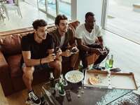 group of three men sitting on a brown leather sofa playing video games and eating snacks and beer