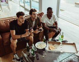 group of three men sitting on a brown leather sofa playing video games and eating snacks and beer