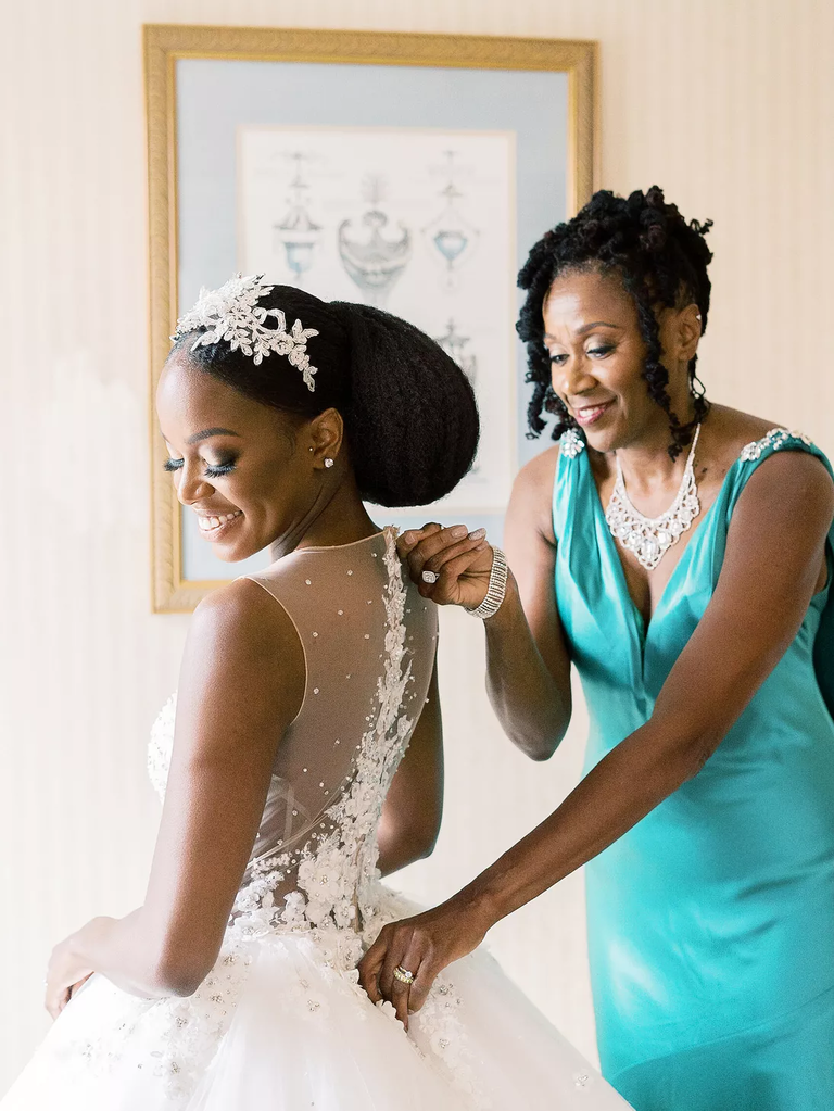 A mother adjusts her daughter's wedding dress as they both smile.