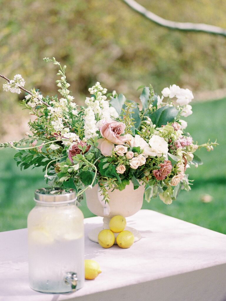 self serve wedding drink station with lemonade in glass beverage dispenser next to pastel pink floral centerpiece