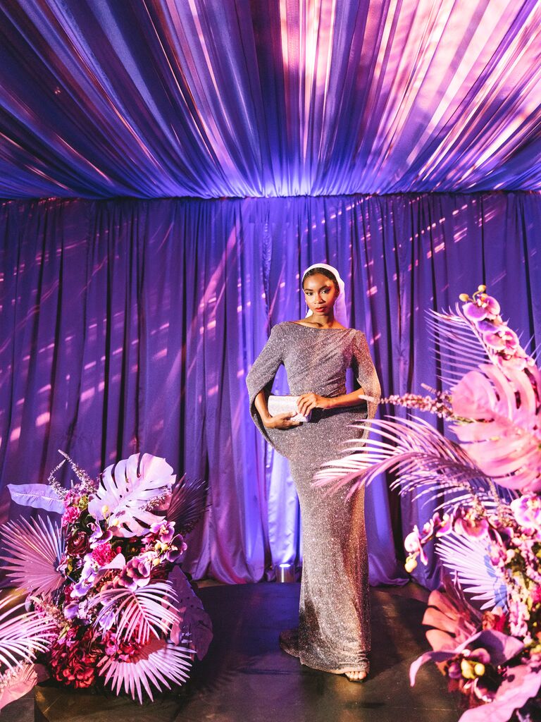 A bride poses for the camera in a stunning silver gown, standing among bright pink monsteras and ferns.
