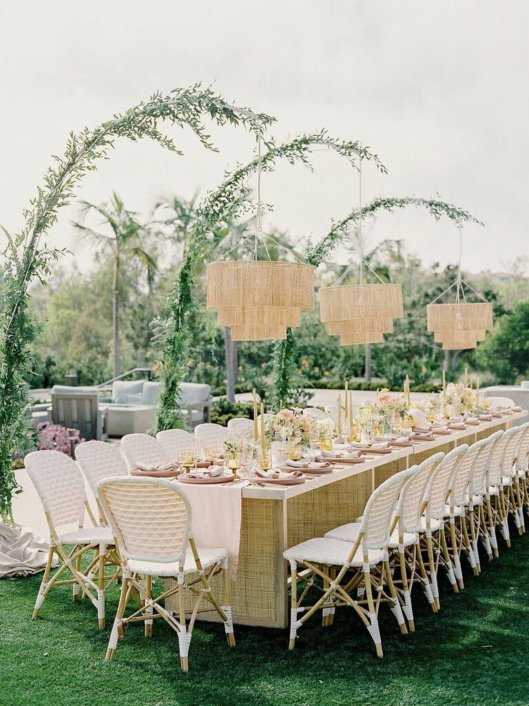 modern outdoor wedding reception long banquet table with white riviera chairs and fringe chandeliers hanging from greenery arches