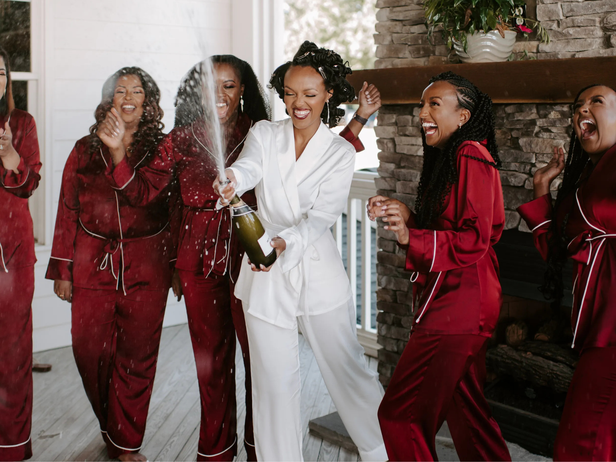 Bride wearing white pajamas and bridesmaids wearing matching red bridesmaid pajamas popping champagne