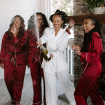 Bride wearing white pajamas and bridesmaids wearing matching red bridesmaid pajamas popping champagne
