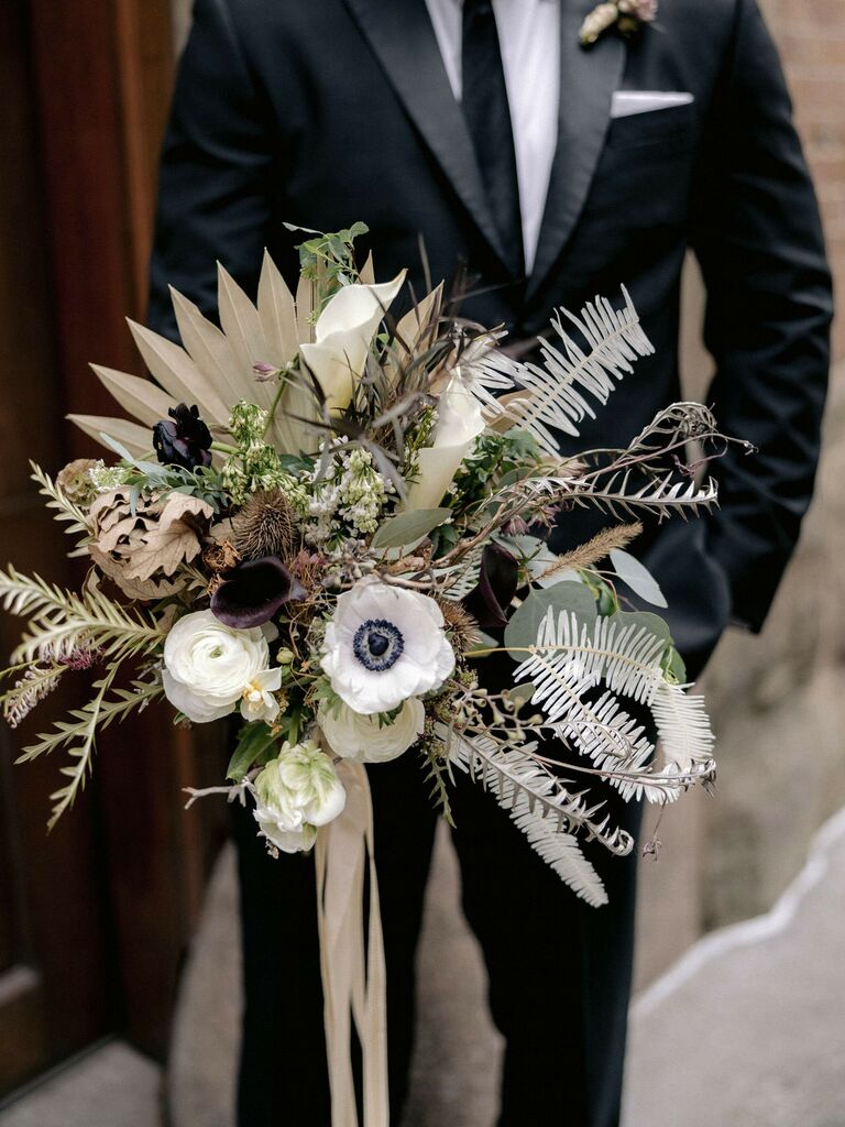 groom holding modern boho winter wedding bouquet with dried fan palm, anemones, ranunculus and greenery in ivory, beige and green tones