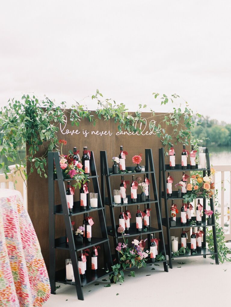outdoor wedding favor display with wine bottles stacked on angled bookcases against backdrop that says "love is never canceled"