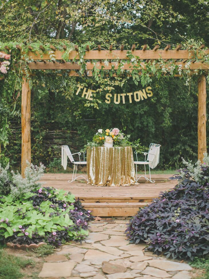 Outdoor sweetheart table under a wooden pergola with a banner hanging above that says the suttons