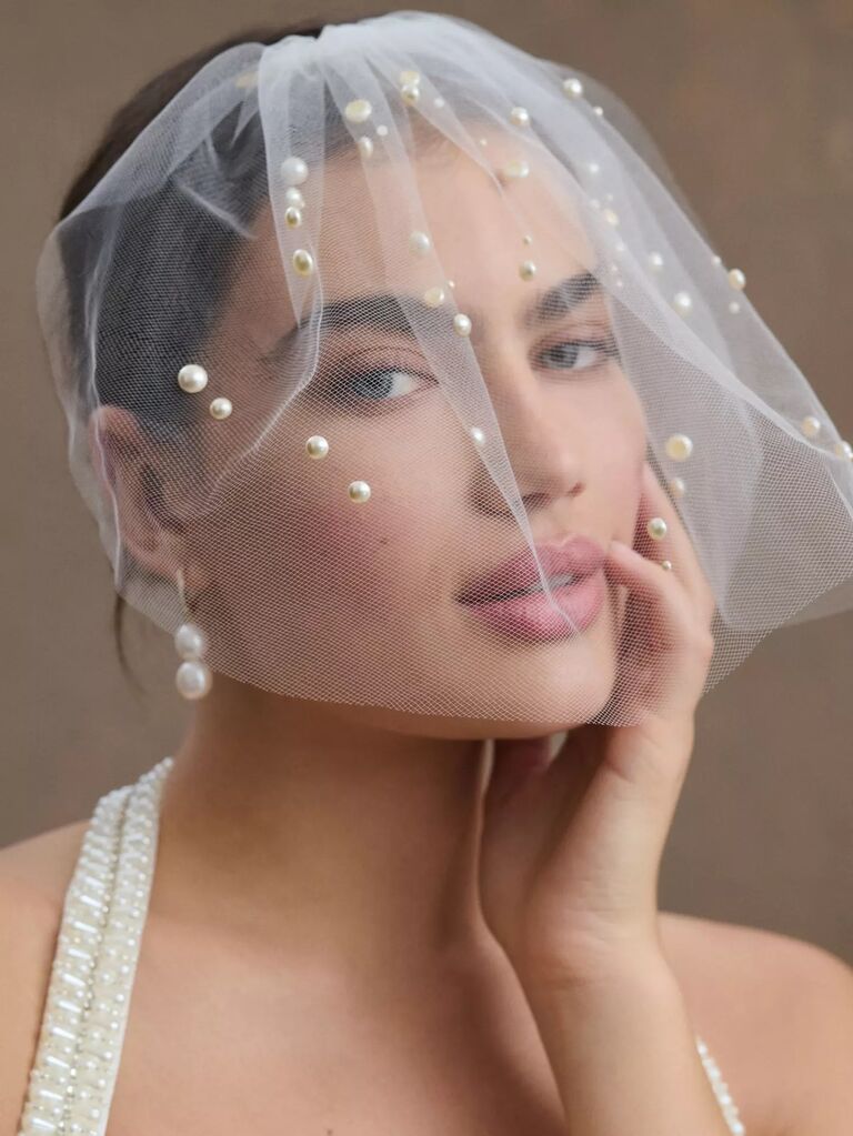 Model wears a short birdcage veil with various pearl embellishments. 