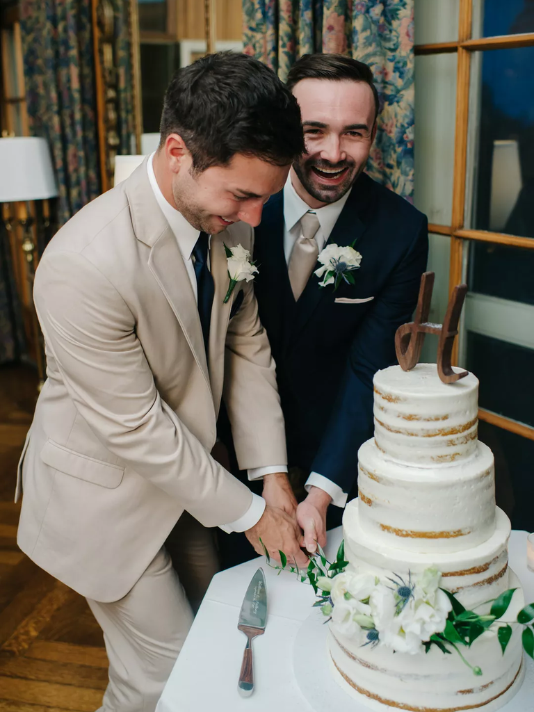 A same-sex couple smiles for the camera while they cut their wedding cake