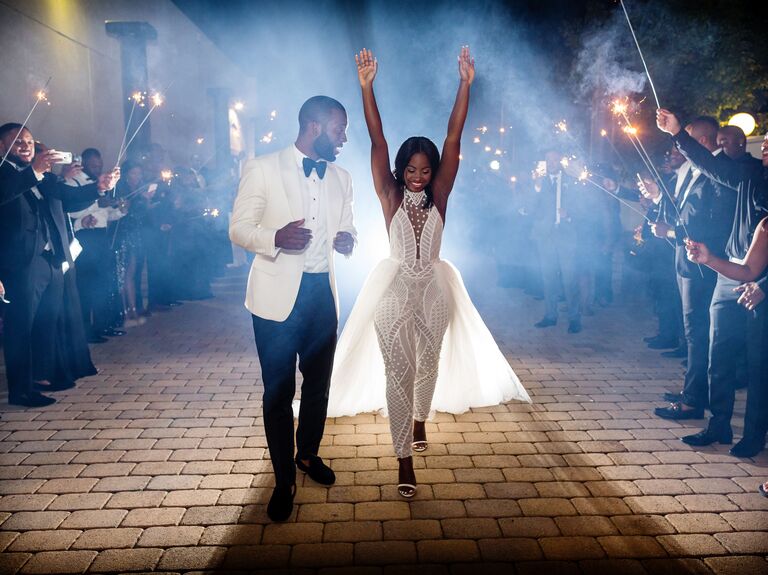 couple walking through sparkler tunnel after wedding wearing white tuxedo and white bridal jumpsuit with tulle train