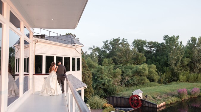 Bride and groom standing together on a balcony overlooking lush gardens and a pond at The Mill Lakeside Manor