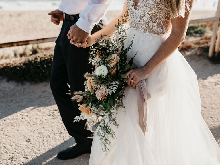 Bride holding wedding flower bouquet walking on beach with groom