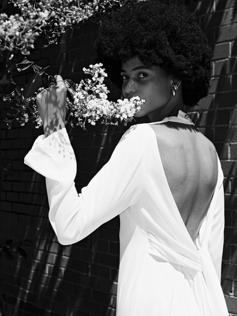 Model wears a white gown with an open back while smelling flowers. 