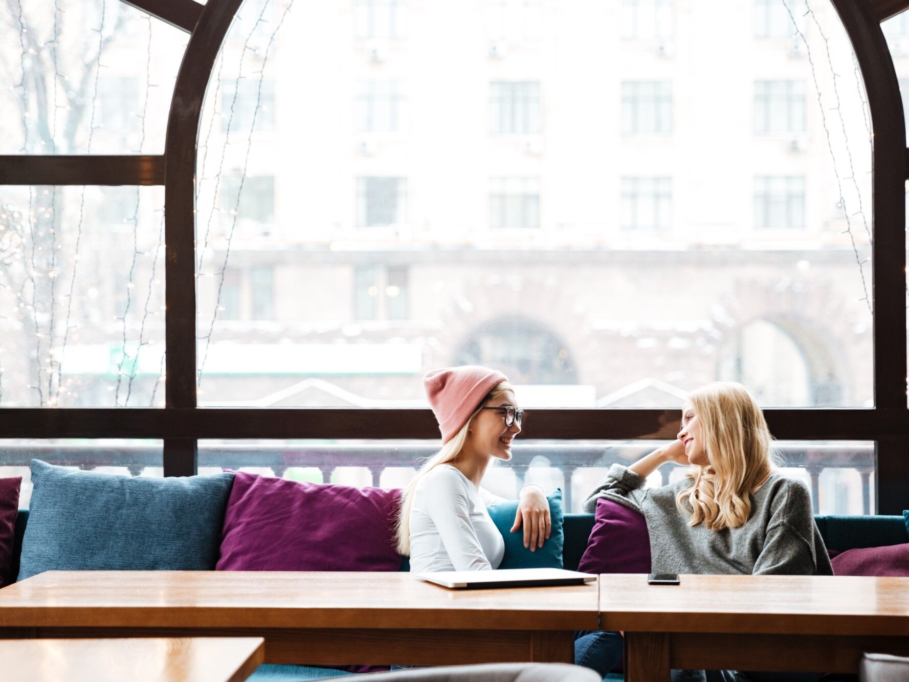 Two women chat while sitting on a couch in front of a large window. 