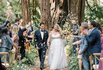 A happy couple walk back down the aisle, beaming, with their friends and towering redwoods in the background.