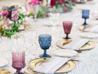 Colorful glassware in burgundy and blue with gold chargers on a lace tablecloth at a wedding reception