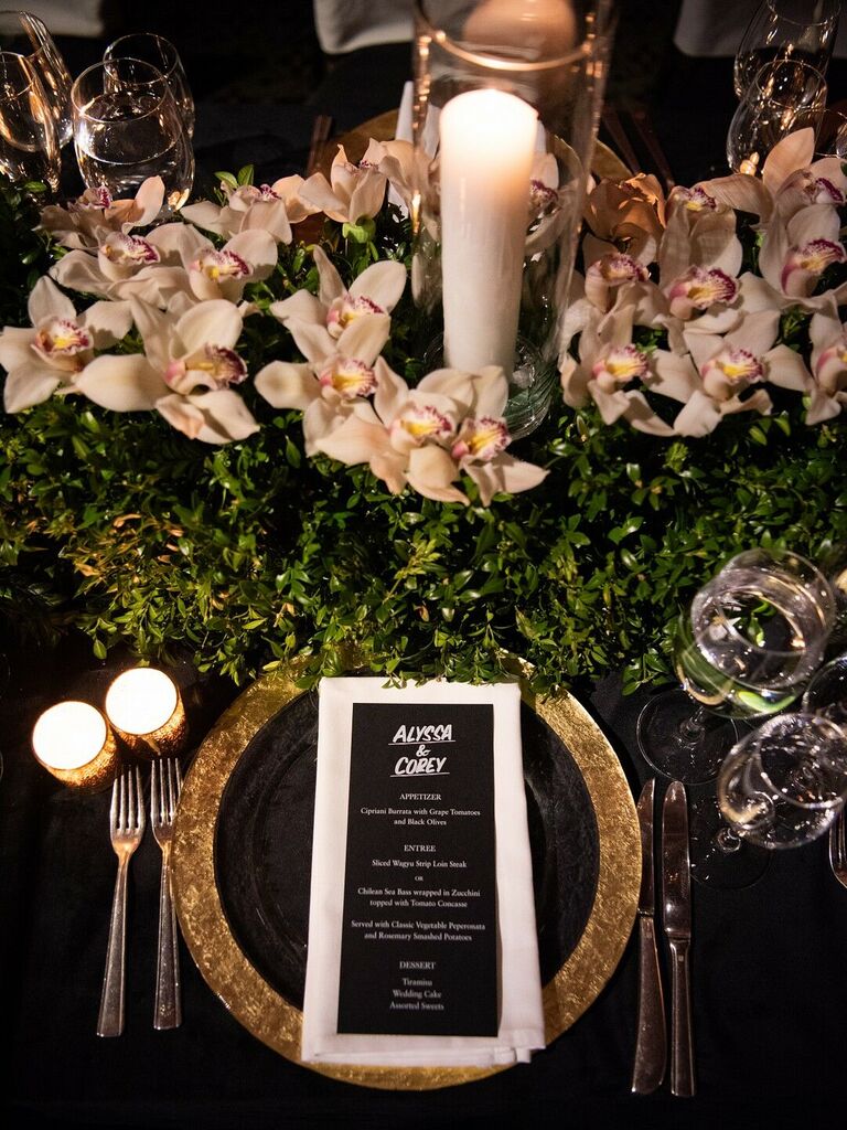 elegant NYE wedding tablescape with white orchids, greenery, white pillar candles and gold charger plates against black tablecloth
