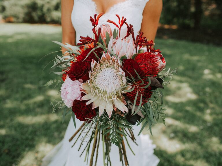 A bride holds a breathtaking bouquet of proteas and peonies in shades of burgundy and pale pink.