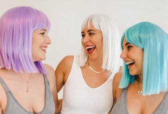 Three friends smile at one another while wearing multicolored wigs