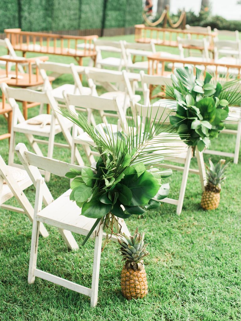 tropical wedding aisle markers with pineapples and monstera leaves tied to white folding chairs