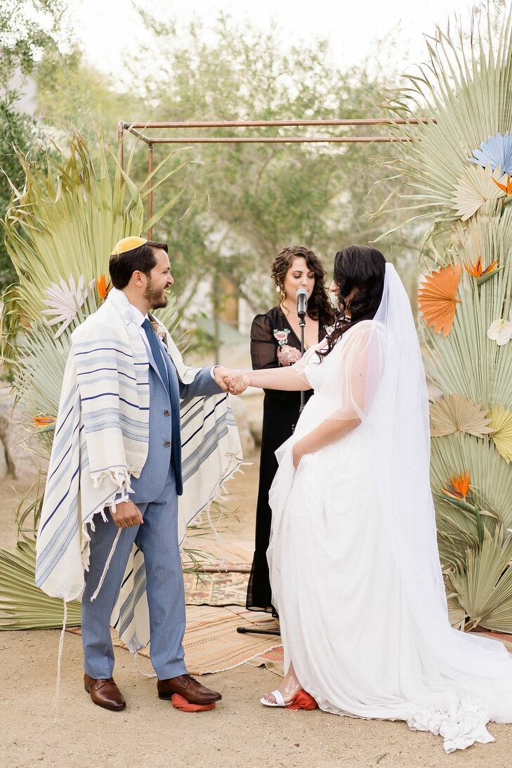 Couple standing in front of copper chuppah decorated with dried palm fronds. 