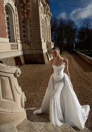 Chantilly lace cascades over the bare shoulders and structured bodice of the Code gown. The strapless neckline meets delicate drapery and exposed boning above a basque waistline. This body-skimming Mikado and lace silhouette balances a low, exposed back with a sweeping train of chapel and court lengths. WONÃ Concept pairs the base with a detachable soft ivory overskirt. Intricate floral embroidery details complete a layered bridal look.