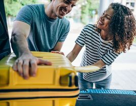 Newlyweds placing yellow hard-shell luggage in trunk 