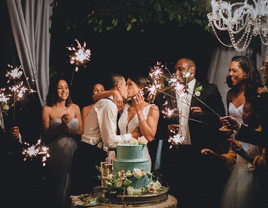 Newlyweds kissing next to wedding cake while guests hold sparklers.
