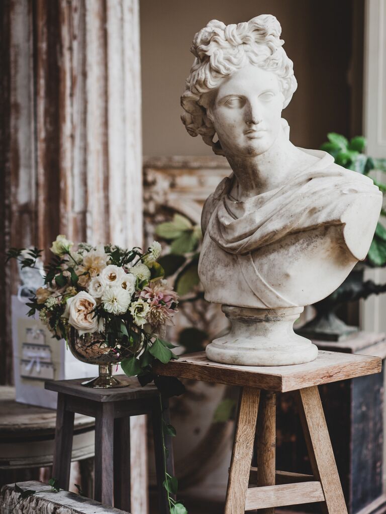 roman goddess statue on a wooden stool next to small white and blush wedding centerpiece