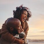 young couple laughing on the beach while man carries woman piggyback 