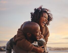 young couple laughing on the beach while man carries woman piggyback 