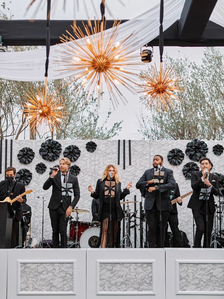 live band performs on stage at luxury wedding with modern sunburst chandeliers and custom flower wall decorating the venue