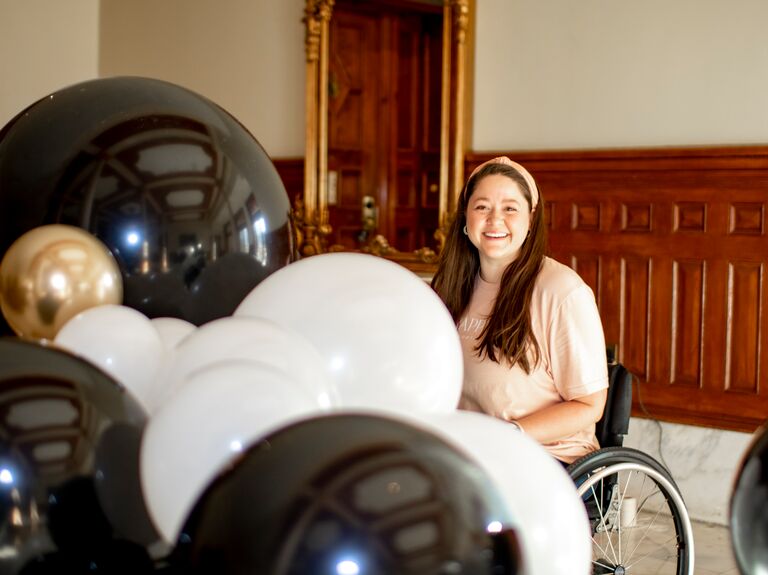A woman in a wheelchair smiles at the camera from where she sits, surrounded by celebration balloons in black, white, and gold.
