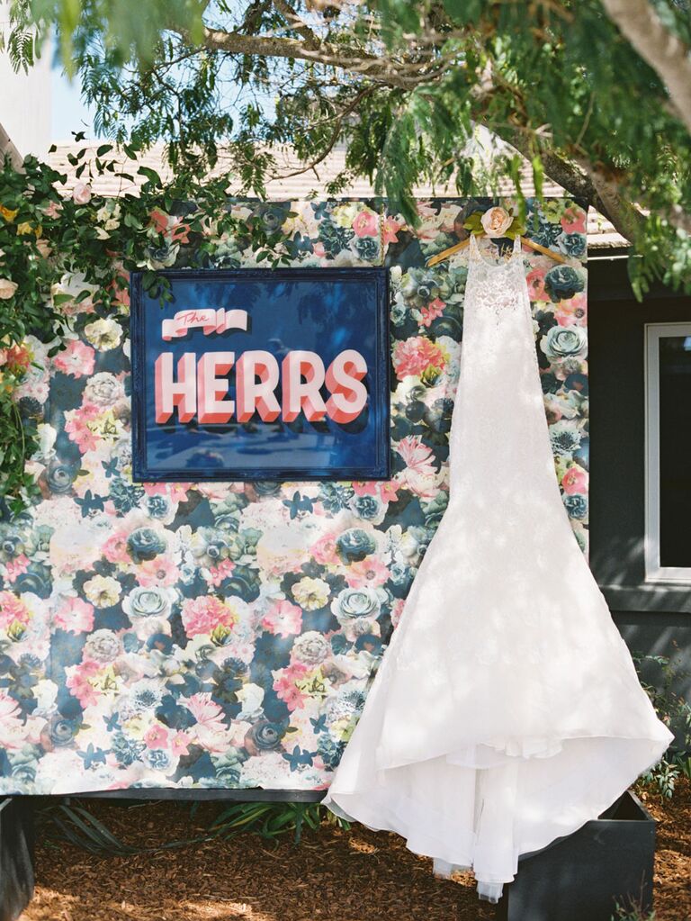 Retro pink custom wedding signage hanging on floral wallpaper next to wedding dress