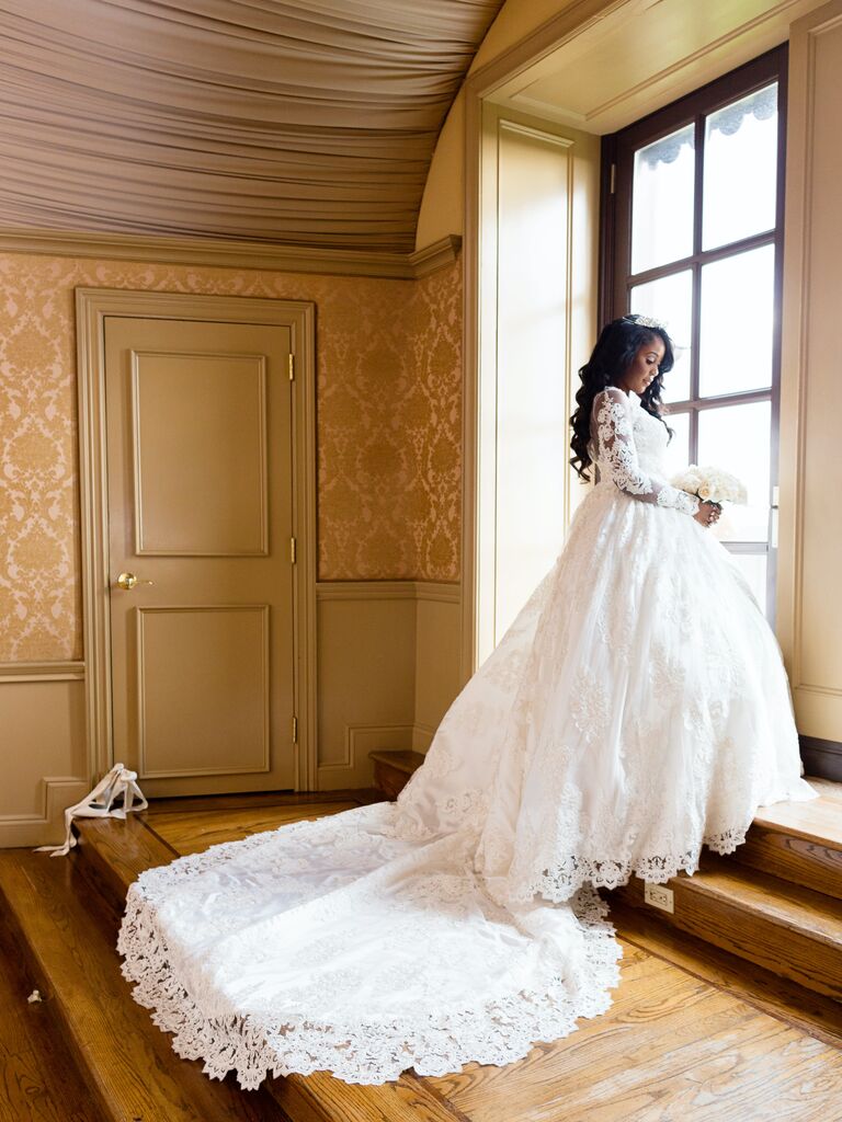 bride wearing lace ball gown wedding dress with cathedral train stands in front of floor length window holding her bouquet
